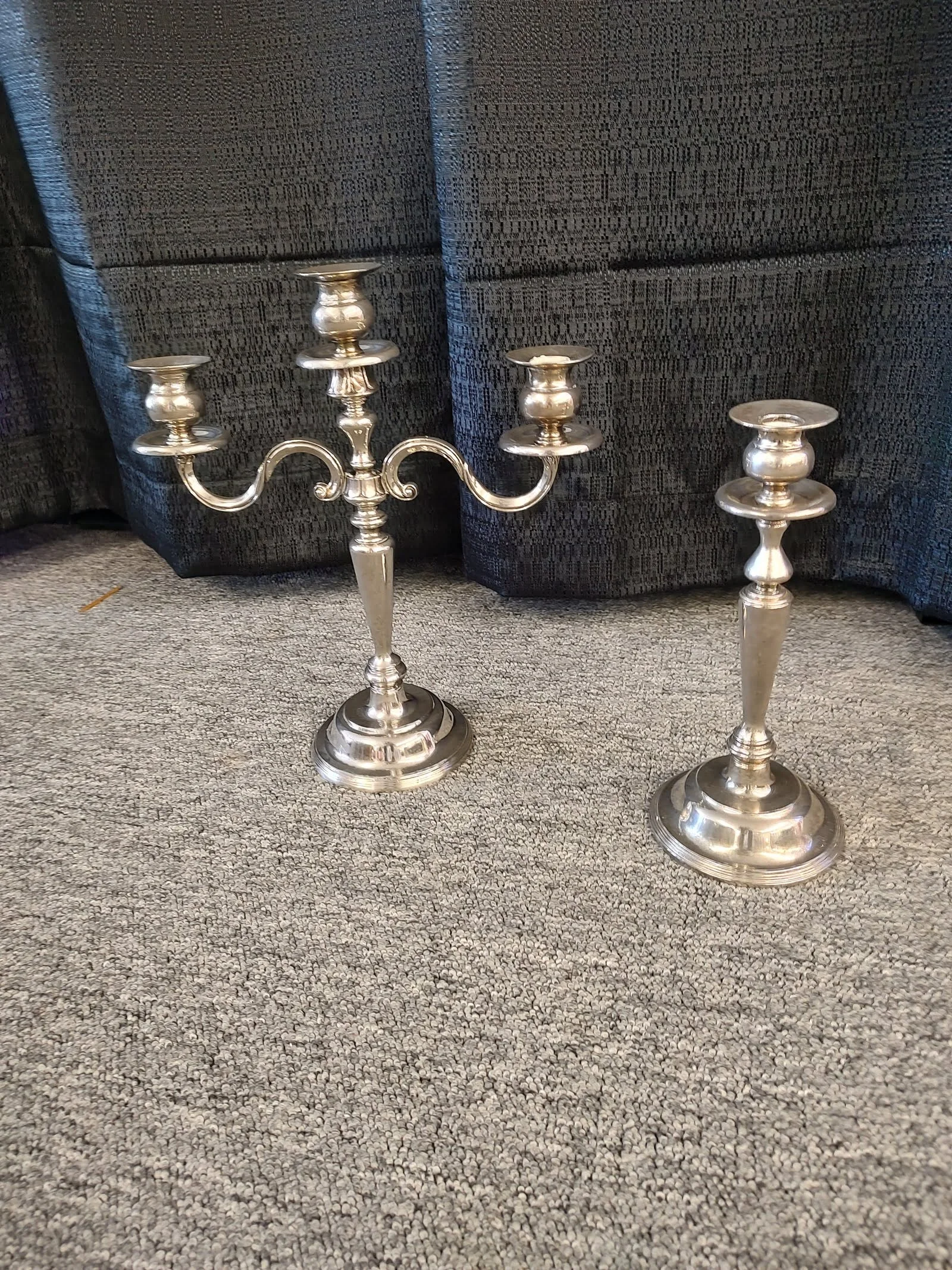 Two silver candle holders on a carpeted floor, one with three arms and one single.