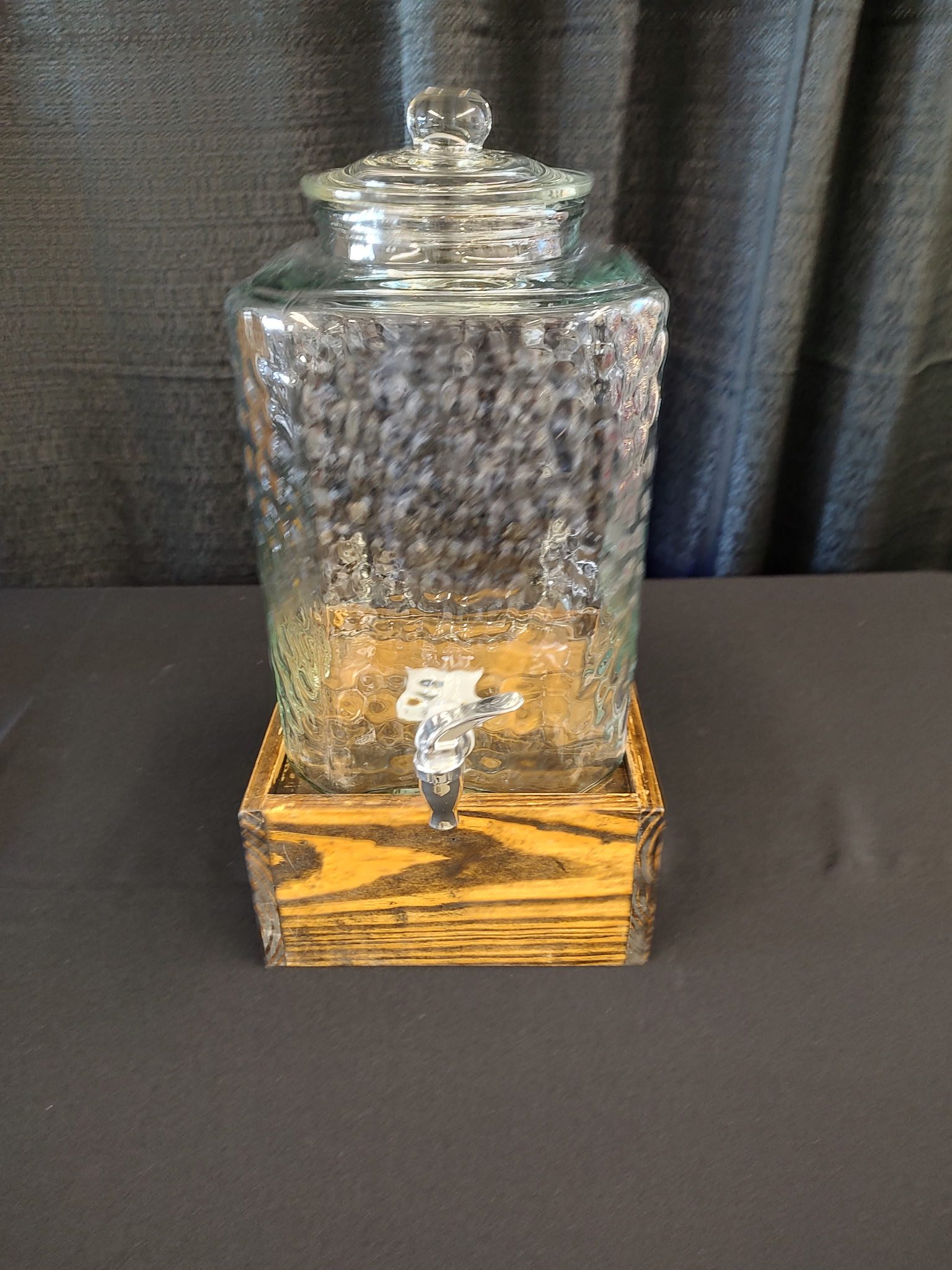 Glass beverage dispenser with wooden base on a black surface.