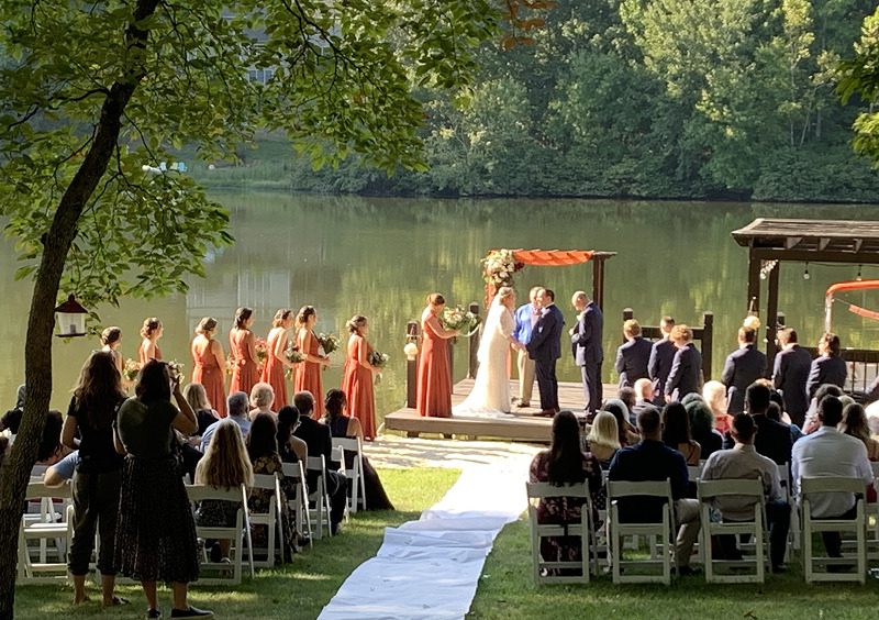 Outdoor wedding ceremony by a lake with guests seated and bridal party standing.